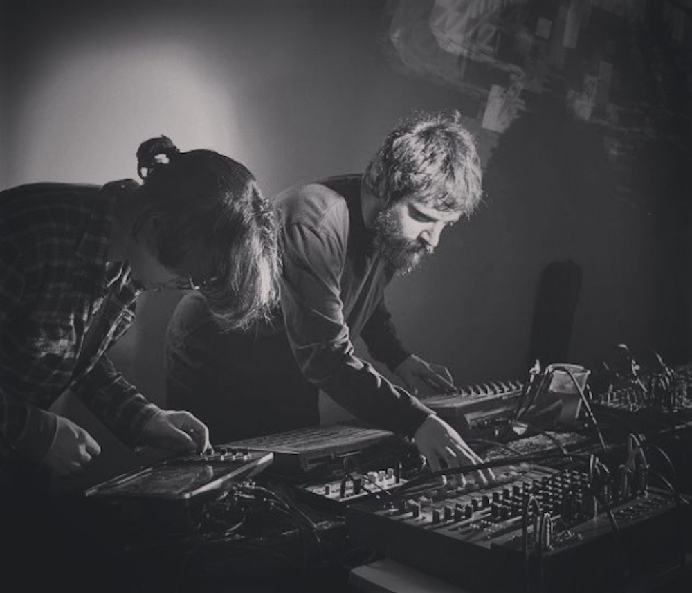 Imagen con dos personas tocando sintetizadores sobre una mesa, con la cabeza mirando hacia abajo